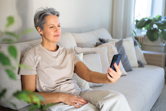Mature beautiful middle-aged woman with gray hair and poor eyesight sits on the couch