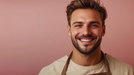 Obraz premium Smiling young man with a beard and wavy brown hair, wearing a beige apron, looking friendly and confident against a soft pink background