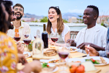 Group of diverse young adult joyful friends barbecue summer party reunion eating on rooftop. Millennial people laughing sitting around table with red wine glasses enjoying free time together outdoors