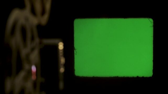 Film reels spin on a cinema projector and are projected as a green screen on the surface.
