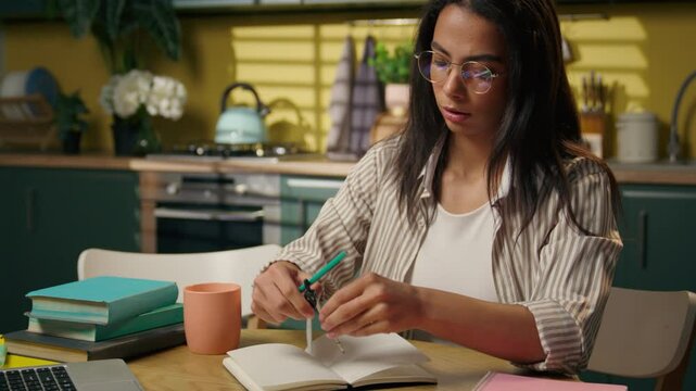 Indian Arabian ethnic woman latina teen girl freelancer home kitchen studying writing notes compass for drawing female high school university student pupil write notebook pencil draft distant studying