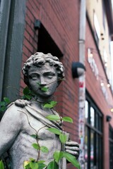 Fototapeta premium Close up of statue covered with climbing green vines on the sidewalk next to a red brick building. 