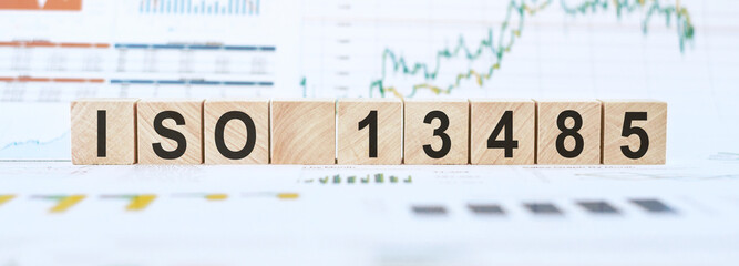Wooden blocks spelling out ISO 13485 standard on a table with financial charts in the background.