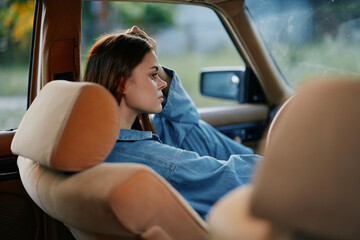 Thoughtful young woman sitting in a vintage car, dressed in casual denim attire, with a pensive expression against a blurred outdoor background.