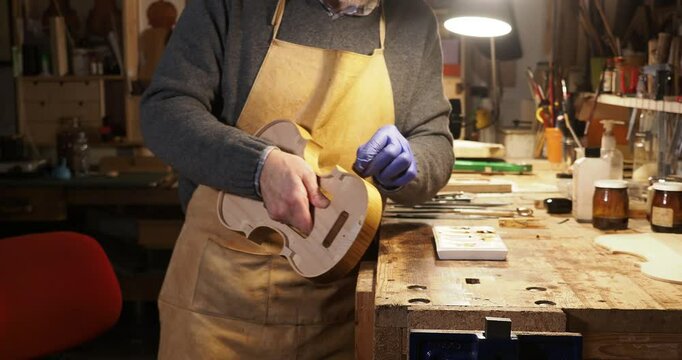 Close up of Luthier wearing an apron and gloves is carefully tampon the ribs of a violin with casein in his workshop, illuminated by a lamp, surrounded by tools and materials , Cremona, Italy