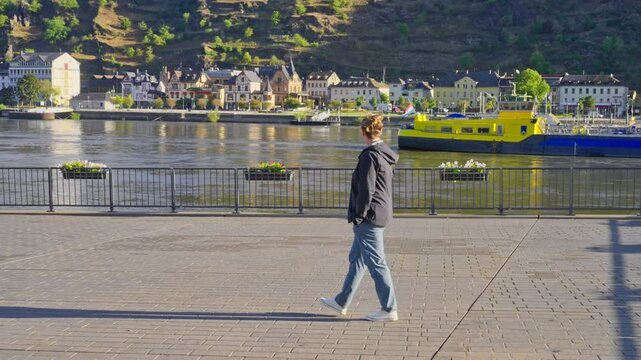 Caucasian adult woman enjoying sunny early morning walk on a riverfront sidewalk with picturesque views of the nearby town, Rhine River Valley, Germany
