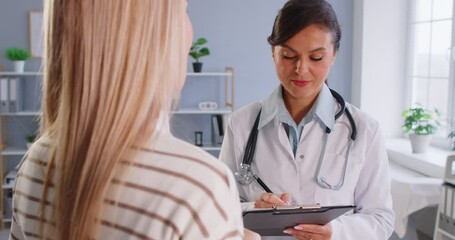 Doctor conducts medical examination of young woman and makes notes in medical card. Female doctor wearing white coat and smiling writes information on clipboard while patient speaks. Medicine concept.