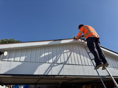 Caucasian man wearing high-visibility clothing up a ladder holding a roll of tape while carrying out a garage roof repair on a concrete block structure in New Zealand on a sunny day