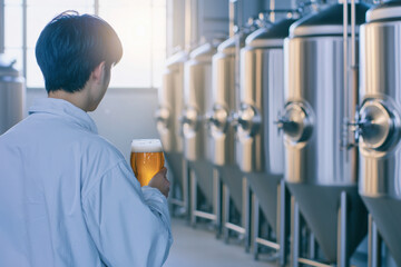 brewer in white coat examines glass of beer in brewery with large fermentation tanks