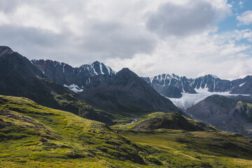 Dramatic scenery in alpine valley with creek among green hills and rocks with view to rocky sharp pointy peak, large snow-capped peaked top, mountain range and big glacier tongue under gray cloudy sky