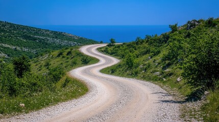 Curved gravel road meanders through lush green hills leading to a serene blue ocean under a bright clear sky.