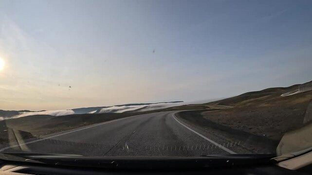 Driving through the tundra in northern Norway on the E69 road. The landscape features plains and hills covered with moss and lichen, with patches of snow. The sun shines directly ahead.
