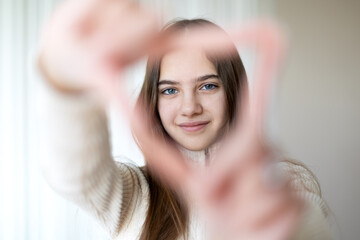 Fototapeta premium Girl framing her face with her hands in a heart shape. A young girl with brown hair smiles gently while framing her face with her hands in a heart shape, standing against a softly lit neutral