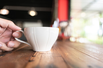 Close-up of a hand holding a white coffee cup on a rustic wooden table in a cozy café setting.