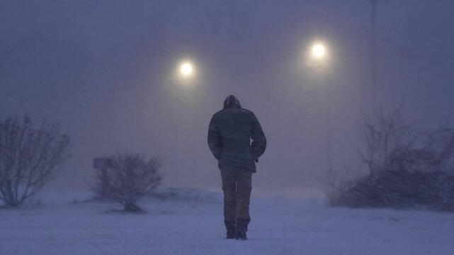 Homeless Man Walks Away from Camera in Snowy Blizzard Towards Streetlights at Night