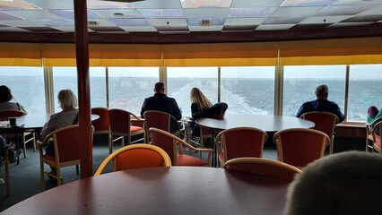 Guests relax inside a boat lounge, watching the calm waves and distant shoreline through large windows. The atmosphere is peaceful despite the cloudy weather.