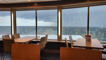 A tranquil dining area features wooden tables and chairs, with large windows overlooking a choppy ocean under cloudy skies. The scene evokes a sense of calm and connection to nature.