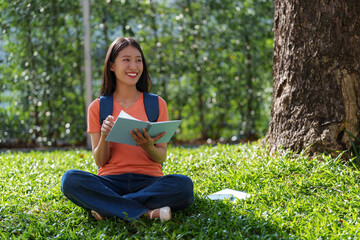 Fototapeta premium Young happy asian female student taking notes in a notebook while sitting under the shade of a tree on a green lawn in a college campus, enjoying the fresh air and the beautiful day