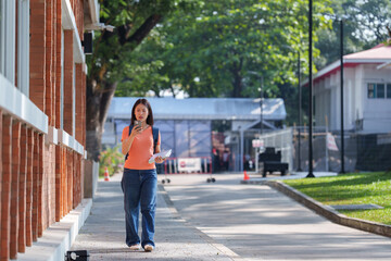 Young asian woman student is walking on a university campus while using her smartphone and carrying books, enjoying the pleasant weather and academic atmosphere
