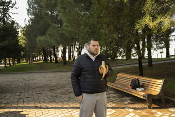Obraz premium Man snacking on healthy snack banana in the city park, eating healthy food in cold weather, casual urban environment with nature.
