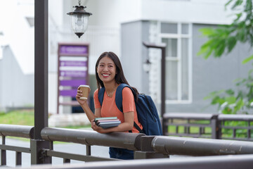 Happy asian female student holding a coffee and books, leaning on a railing on a university campus...