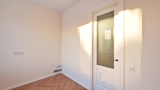 Bright, minimalist room with parquet flooring. Natural light through the window and glass paneled door. Cream walls, simple decor.