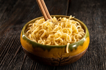 Steaming hot noodles being lifted from a  bowl by a pair of wooden chopsticks. The golden-yellow noodles cascade down, releasing wisps of steam that curl. Instant noodles on dark background. Food conc