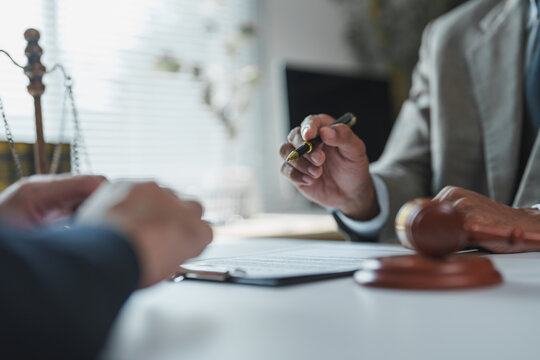 Lawyer holding a pen while explaining contract details to a client in a law firm or notary office, with a gavel and scales of justice prominently displayed on the desk