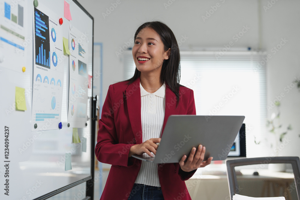 Poster smiling businesswoman working on a laptop while presenting marketing data and analysis on a whiteboa