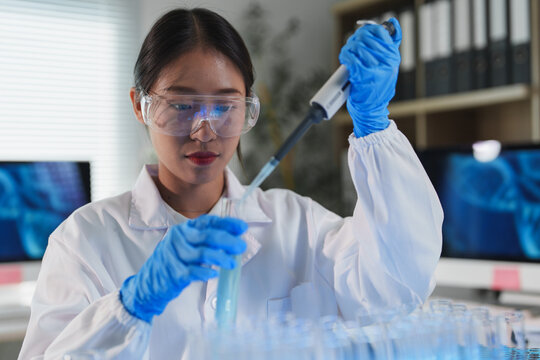 Young female scientist wearing protective glasses and gloves using micropipette adding reagent into test tube for research in modern laboratory, with test tubes and computer in background