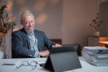Happy mature business executive working late in an office, using a digital tablet while surrounded by paperwork and a keyboard, embodying professionalism and dedication