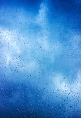 Rain drops on the glass window surface, blue clouds visible through the rain, day time shot. Rainy background, vertical orientation