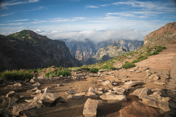 Vast rocky terrain stretches across the foreground, leading to towering mountains shrouded in clouds. Sunlight illuminates the landscape, highlighting its natural beauty.