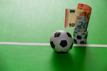 A close-up of cups featuring soccer ball designs, accompanied by ten and twenty-euro banknotes on a green background near a white line with a soccer ball, representing sports betting,