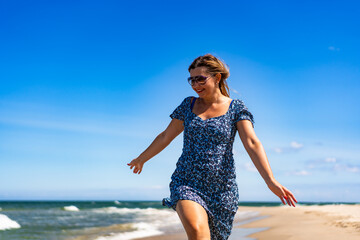 Summertime by sea. Happy middle-aged woman walking on sandy beach on sunny summer day. Front view