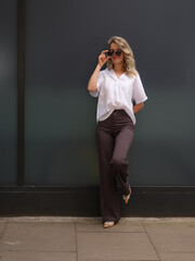 A fashionable woman in a white blouse and dark trousers poses confidently against a modern urban...