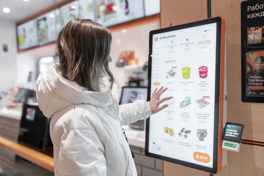 A Customer Interacting with a Digital Menu Kiosk in a FastFood Restaurant Setting. A 30 - to 35-year-old woman in a white winter jacket orders dessert at a fast food restaurant or pizzeria coffee shop