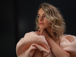 Fototapeta premium A stylish woman in a pink dress with puffed sleeves gazes off-camera with a poised expression, exuding confidence and sophistication against a dark background