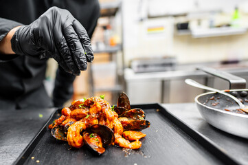 A chef in a black glove garnishes a plate of shrimp and mussels in a vibrant sauce. The stylish dish is presented on a sleek black tray, showcasing culinary artistry in a modern kitchen.