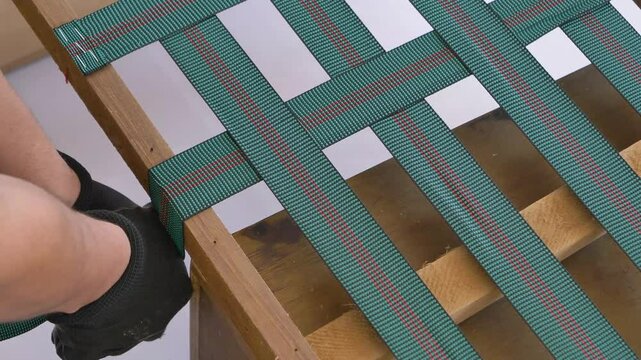 Close-up video showing a worker stretching a strap on a wooden furniture frame during the upholstery preparation process. The strap is being carefully aligned for optimal tension