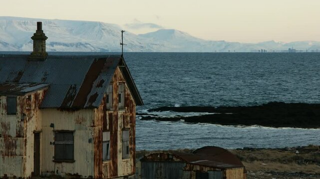 Steady winter shot of degraded house feature snowy mountains, calm ocean at dusk - 4k