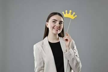Smiling businesswoman holding stick with paper crown on grey background
