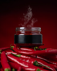 Steaming chili sauce in glass jar surrounded by red hot chili peppers on red background. Cayenne Pepper. Spicy food. Tabasco. Salsa. Piri piri. Ajika. Aji Molido. Harissa. Gochujang. Sambal.