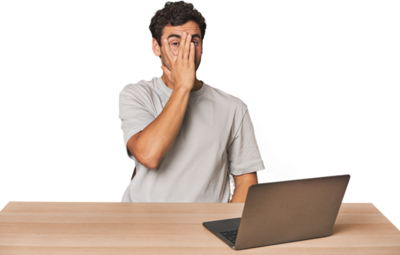 Hispanic young man working on laptop blink at the camera through fingers, embarrassed covering face.