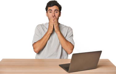 Hispanic young man working on laptop shocked, covering mouth with hands, anxious to discover something new.