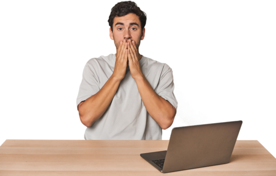 Hispanic young man working on laptop shocked, covering mouth with hands, anxious to discover something new.