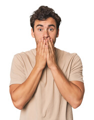 Young Hispanic man in studio shocked, covering mouth with hands, anxious to discover something new.