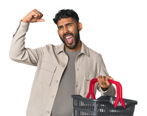 Hispanic male holding shopping basket in studio raising fist after a victory, winner concept.
