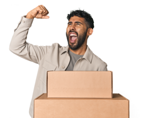 Hispanic male holding cardboard boxes in studio raising fist after a victory, winner concept.