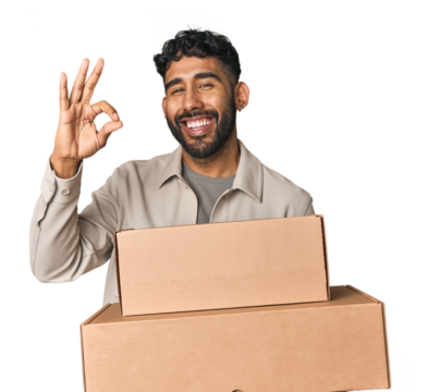 Hispanic male holding cardboard boxes in studio cheerful and confident showing ok gesture.